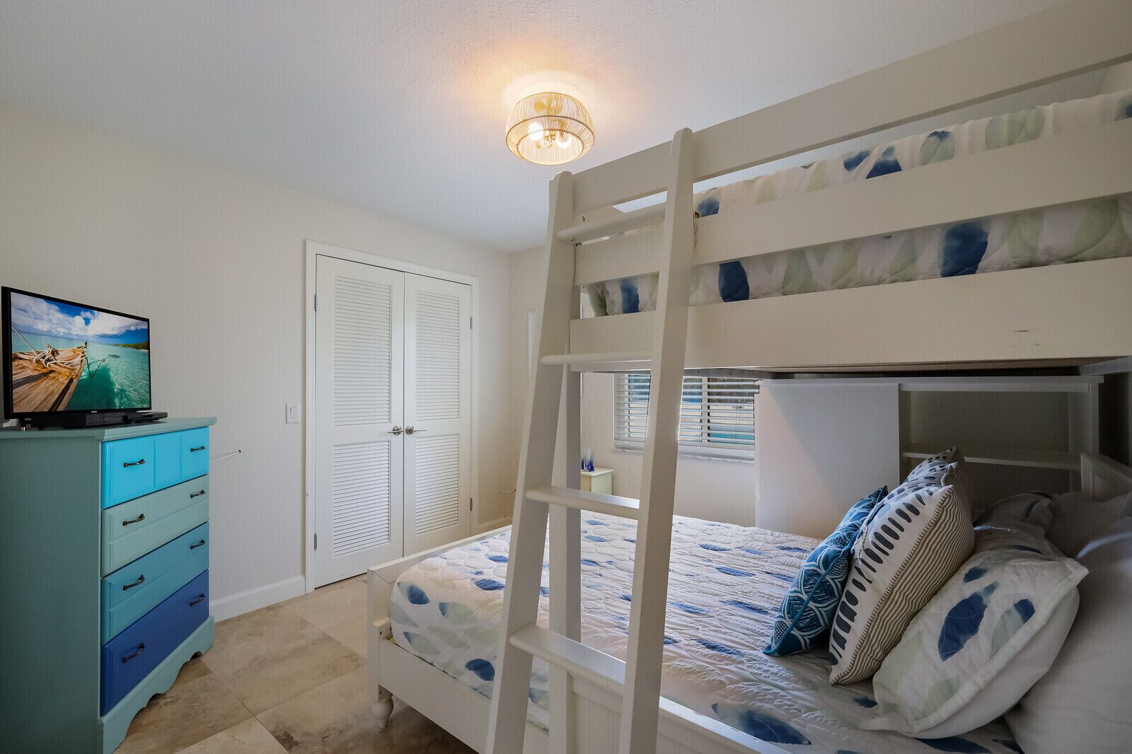 Una cama litera con escalera, un televisor sobre un aparador y una puerta con lámpara.