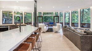 Een moderne keuken met een groot eiland, barstoelen en een inbouwoven. Er is een zithoek met een bank en stoelen, en grote ramen met uitzicht op het buitengebied.