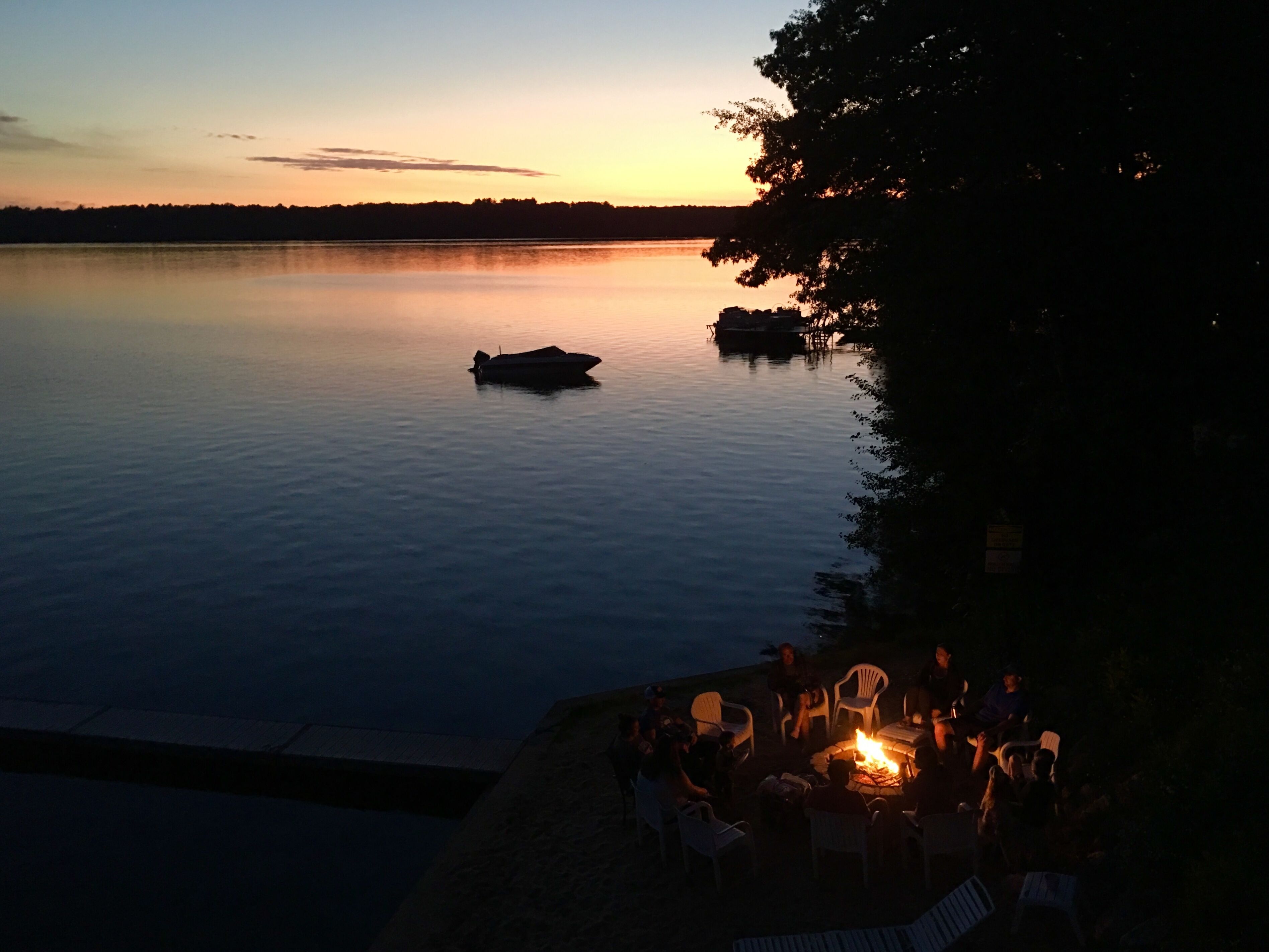 A lakeside view at dusk with a boat on the water and people gathered around a campfire.