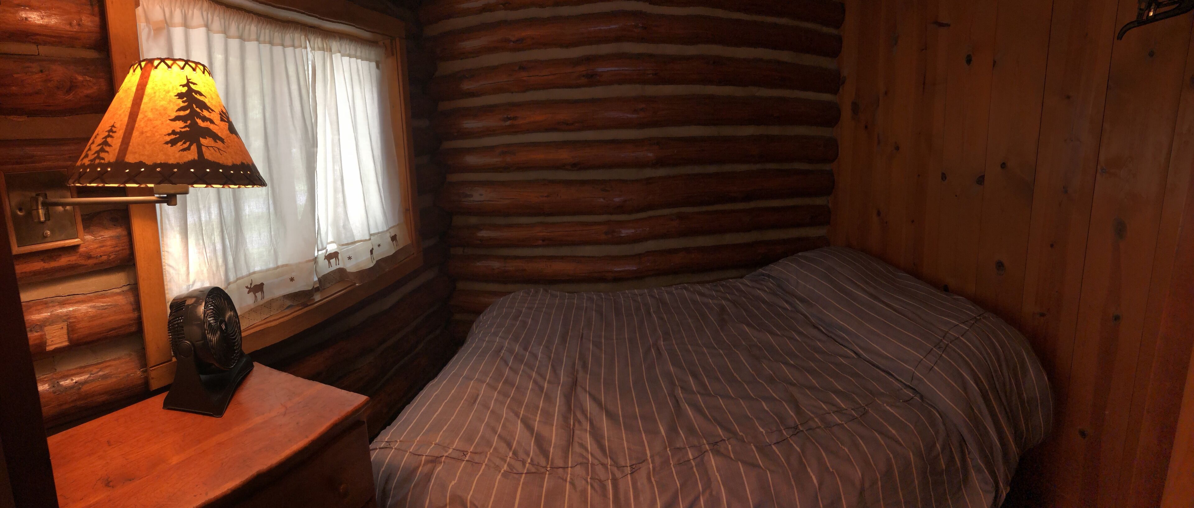 A log cabin room with a bed, a nightstand, a lamp, and a window with curtains.