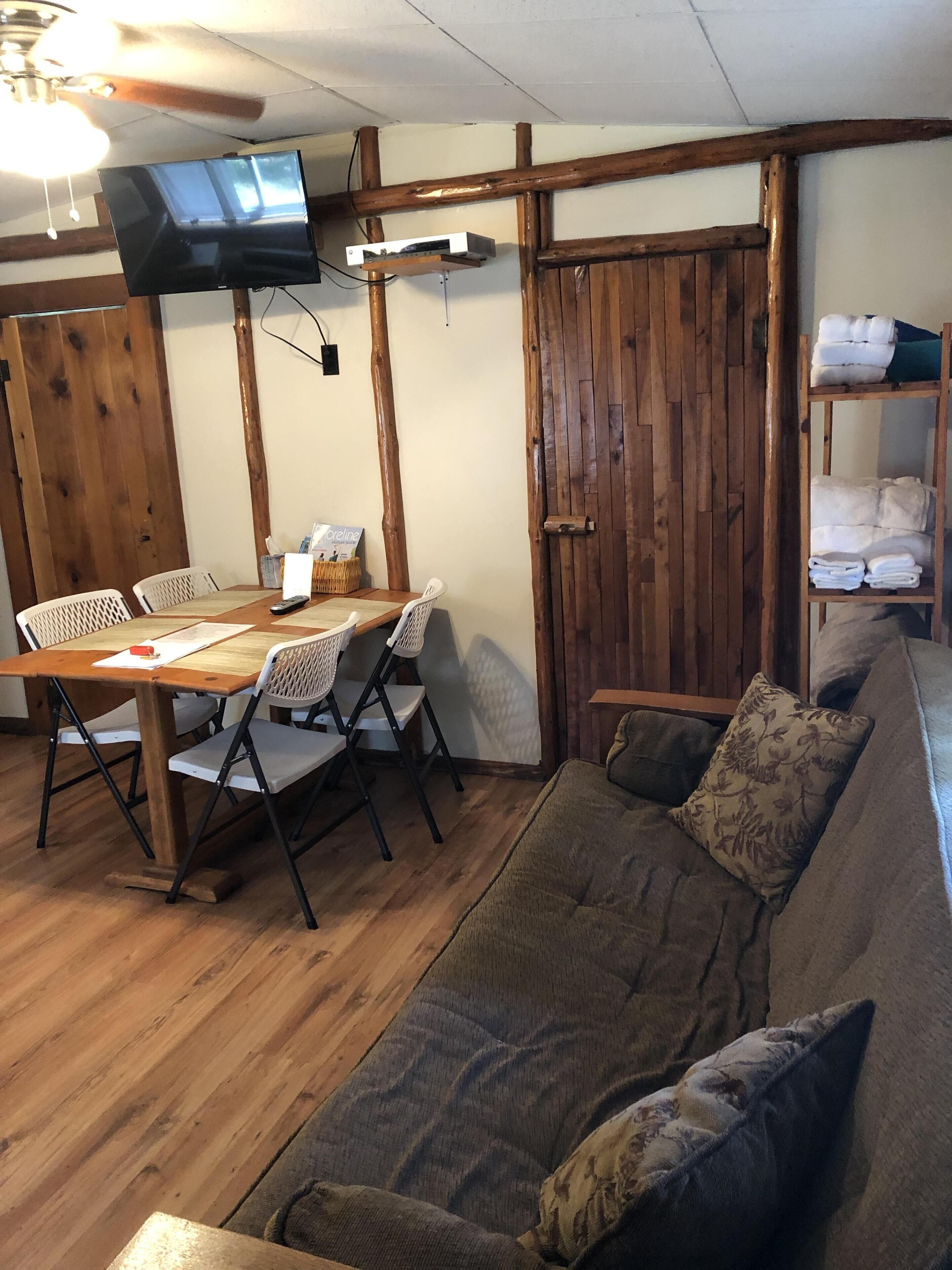 A cozy room with a wooden dining table, chairs, a sofa, a TV mounted on the wall, and a wooden door.
