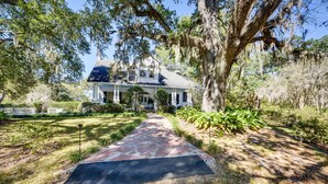 A large house with a porch, surrounded by trees and a stone pathway.