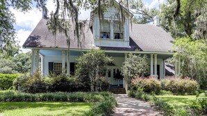 A two-story house with a porch, surrounded by lush greenery and trees.
