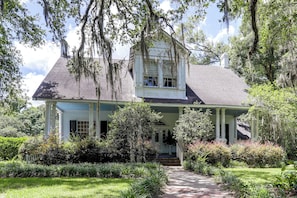 A two-story house with a porch, surrounded by lush greenery and trees.