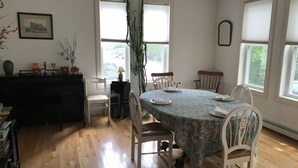 A dining area with a round table, white chairs, and a floral tablecloth. There are two windows with blinds, a potted plant, and a framed picture on the wall.