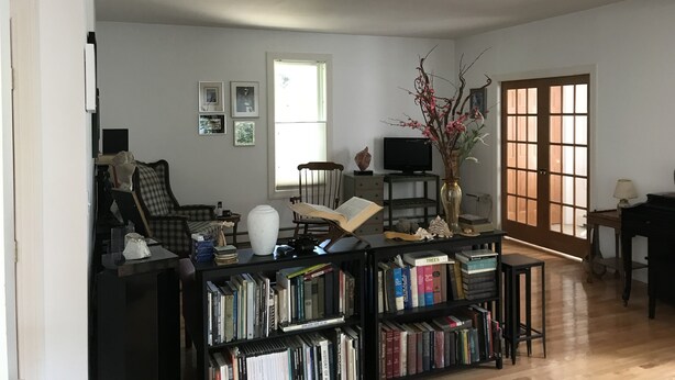 A spacious living room with a large bookshelf, a comfortable armchair, a desk with a computer, and a window providing natural light.