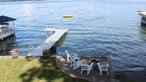 A dock extending into a calm lake with a small boat, a grassy area with Adirondack chairs, and a wooden deck with a covered seating area.