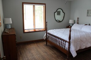 A bedroom with a wooden bed, a window with blinds, a bedside table with a lamp, and a wall-mounted mirror.