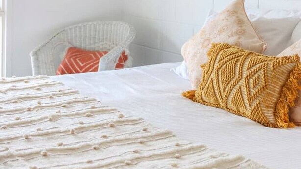 Een slaapkamer met een witte baksteenmuur, een bed met witte lakens, een rieten stoel en een ingelijst schilderij aan de muur.