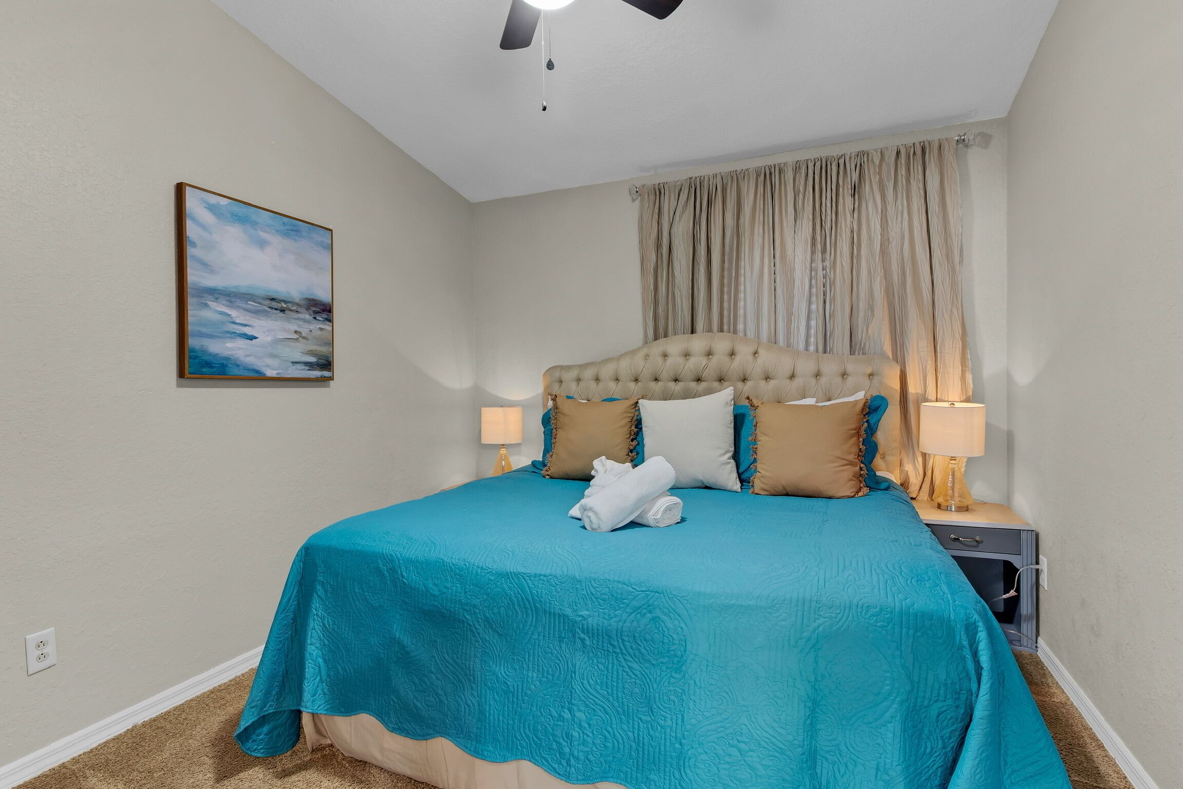Une chambre à coucher avec un grand lit, un ventilateur de plafond, un tableau au mur et une table de chevet avec des lampes.