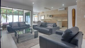 A modern living room with a glass coffee table, grey sofas, and a kitchen area in the background.