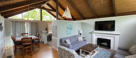 A spacious living area with a high ceiling, wooden beams, a fireplace, a television, and a dining area with a table and chairs.