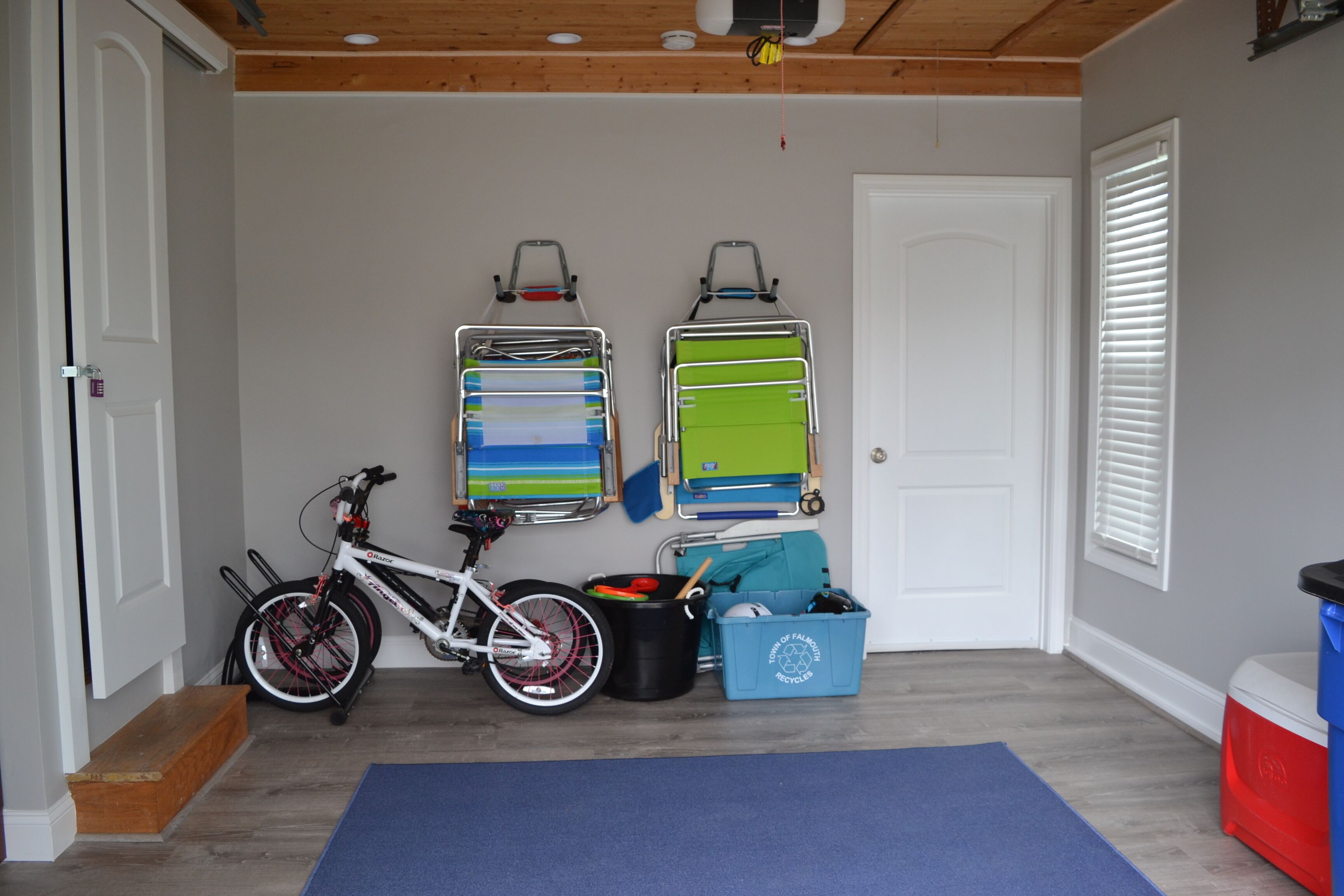 A room with a bicycle, two wall-mounted storage racks, a blue mat, a trash bin, and a cooler.