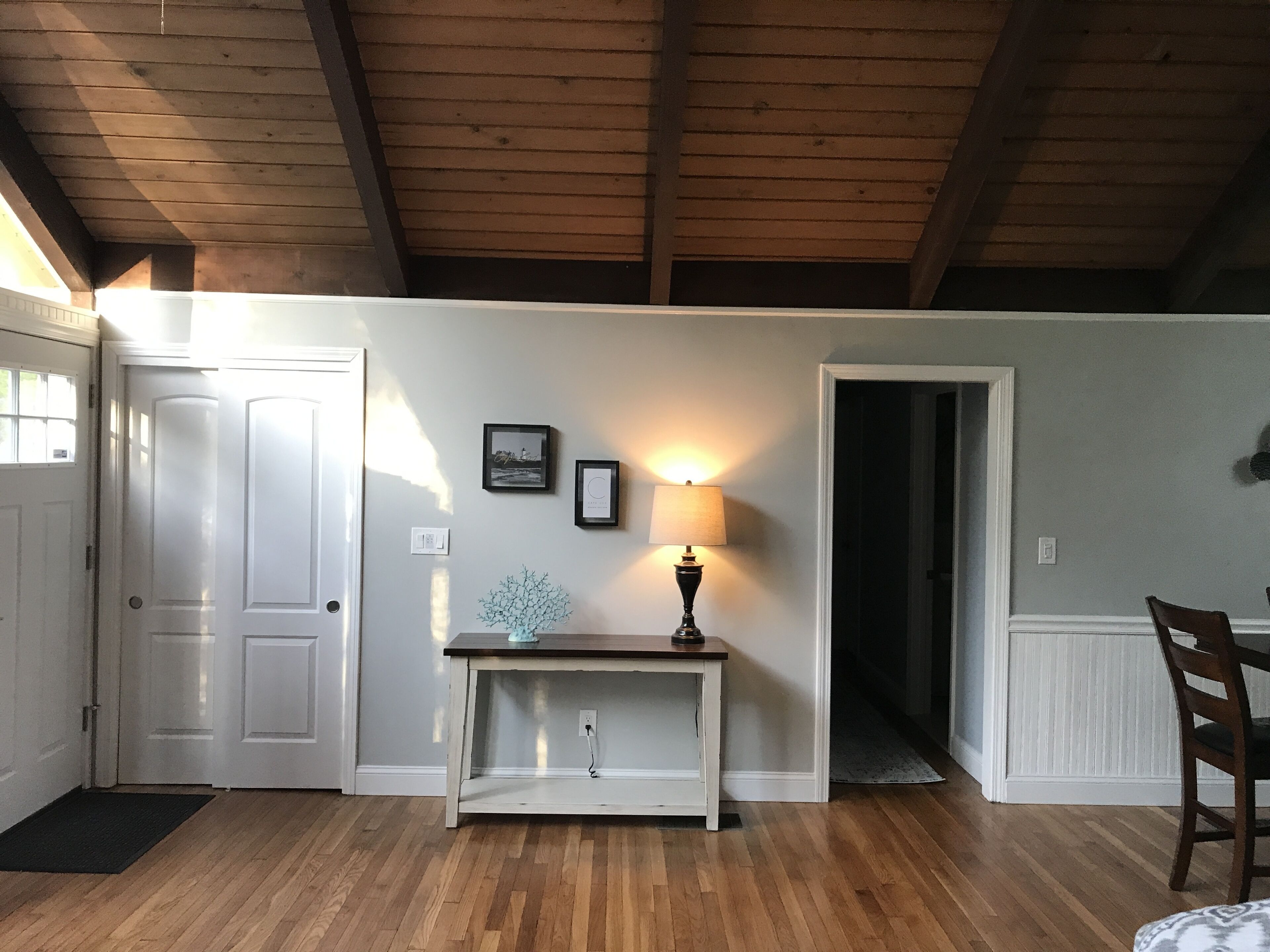 Une pièce avec un plafond en bois, des murs blancs, une porte, une table avec une lampe et une chaise.