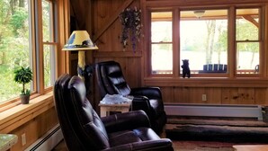 A cozy cabin interior with wooden walls, two leather chairs, a small table, a lamp, and a view of the outdoors through large windows. - Beautiful, Peaceful Lakefront Home in woods. Canoe, Kayaks, ATV trail access. (Sangerville)