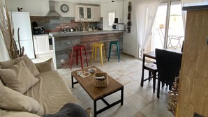 A spacious living area with a sofa, a wooden coffee table, and a dining table with chairs. A kitchen with white cabinets and a built-in microwave is visible in the background.