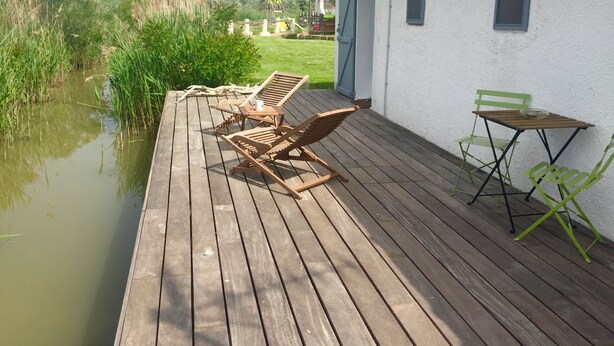A wooden deck with lounge chairs and a table, adjacent to a white building with a tiled roof.