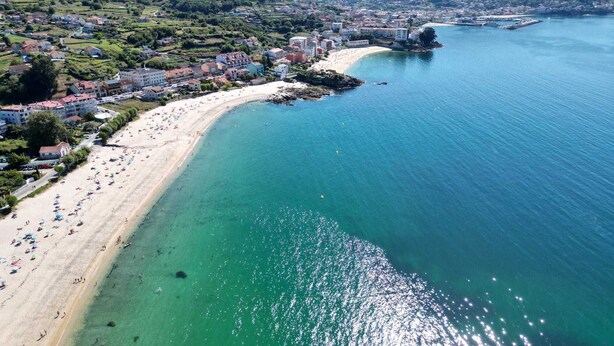 Vista aérea de un pueblo costero con una playa de arena, aguas azul cristalino y una combinación de edificios residenciales y comerciales.