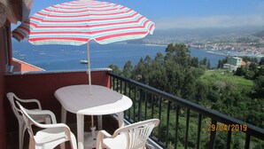 Un balcón con mesa y sillas, un parasol a rayas y vista al mar y al pueblo.