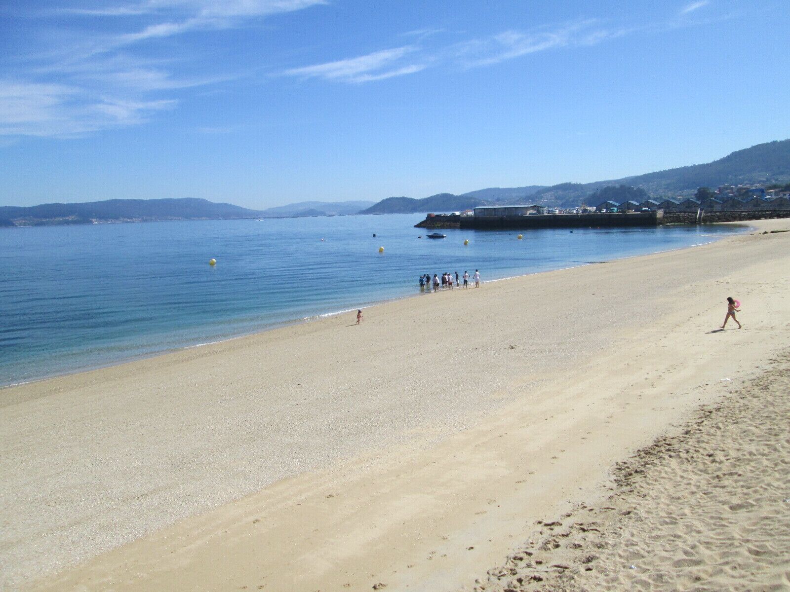 Una spiaggia sabbiosa con acque cristalline blu, alcune persone e colline in lontananza.