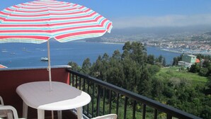 Un balcón con mesa y sillas, un parasol a rayas y vista al mar y al pueblo.