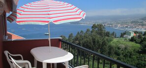 Un balcón con mesa y sillas, un parasol a rayas y vista al mar y al pueblo.