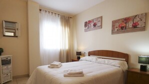 A neatly made bed with white linens and two folded towels, a bedside table with a lamp, and a wall clock.