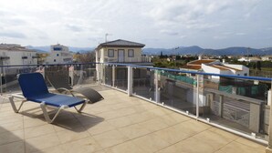A balcony with a blue chair and a sun lounger, a glass railing, and a view of buildings and mountains in the background.