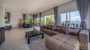 Un vaste espace de vie comprenant un grand canapé d’angle, une table basse et un ensemble de salle à manger. On y trouve également un ventilateur de plafond, de grandes fenêtres avec des rideaux et une vue sur l’océan.