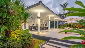 Un espace patio extérieur moderne, doté d’un coin salon abrité, entouré d’une végétation tropicale luxuriante et de palmiers.