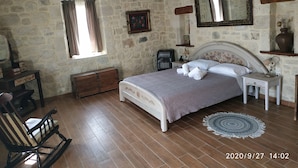 Een rustieke slaapkamer met een stenen muur, houten vloer, een bed met een bloemistisch hoofdbord, een schommelstoel en een plafondventilator.