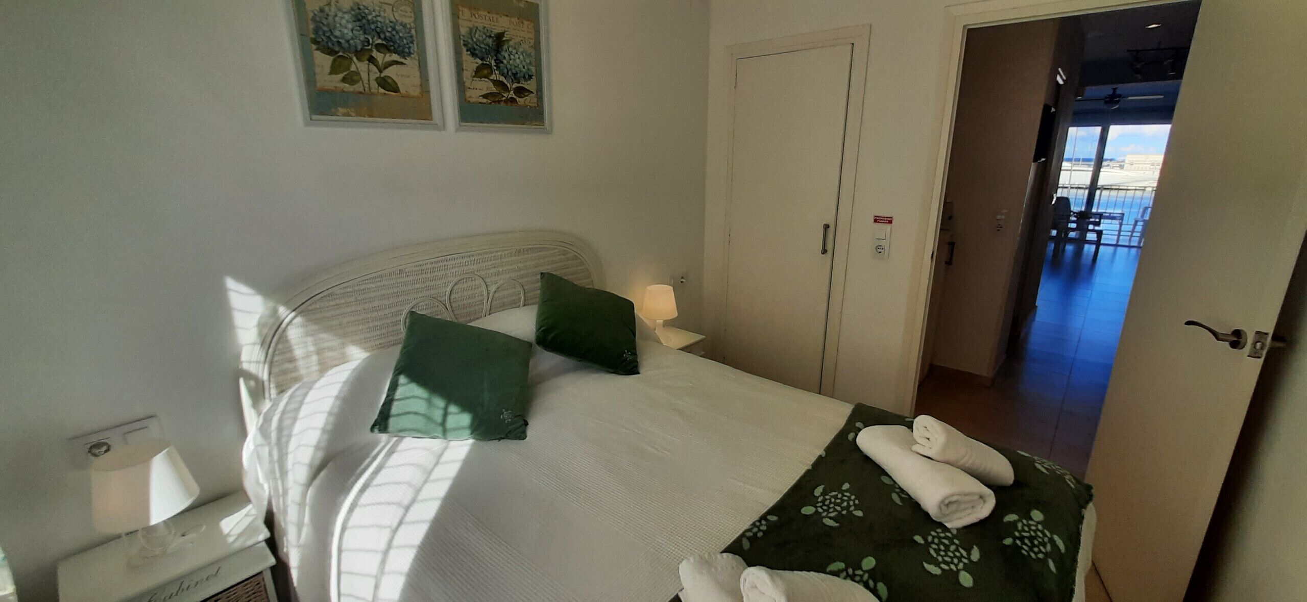 Una cama bien hecha con almohadillas verdes, un cabecero blanco y una manta verde estampada. Hay un juego de toallas sobre la cama y una mesita de noche con una lámpara y una caja de pañuelos.