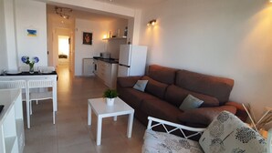 Un salon avec un canapé marron, une table basse blanche et un téléviseur. Il y a également un coin cuisine équipé d’un réfrigérateur et d’un four à micro-ondes.