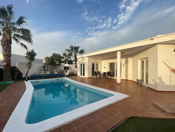 A modern outdoor pool area with a clear blue pool, surrounded by palm trees and a covered patio with a table and chairs.