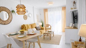Una sala de estar moderna con sofá, mesa de comedor y sillas. Hay un televisor montado en la pared y una lámpara sobre una mesita.