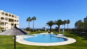 Zona de piscina con vista despejada al océano, rodeada de palmeras y un edificio con balcones.