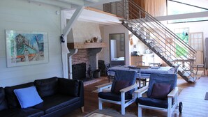 A spacious living area with a black leather sofa, a white armchair, and a dining table. A brick fireplace and a staircase with a metal railing are visible. - Typical regional house, fully and tastefully renovated with old bricks and beams, very sunny surrounded by old trees & vinyards, close to huge beaches & golf courses. (Messanges)