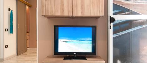 A modern living room with a flat-screen TV on a wooden stand, a small shelf with decorative items, and a view of the ocean through a glass door.