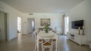 Un grand espace dédié à la salle à manger, avec une table et des chaises blanches, un vase de fleurs, un tableau accroché au mur et un téléviseur posé sur un meuble blanc.