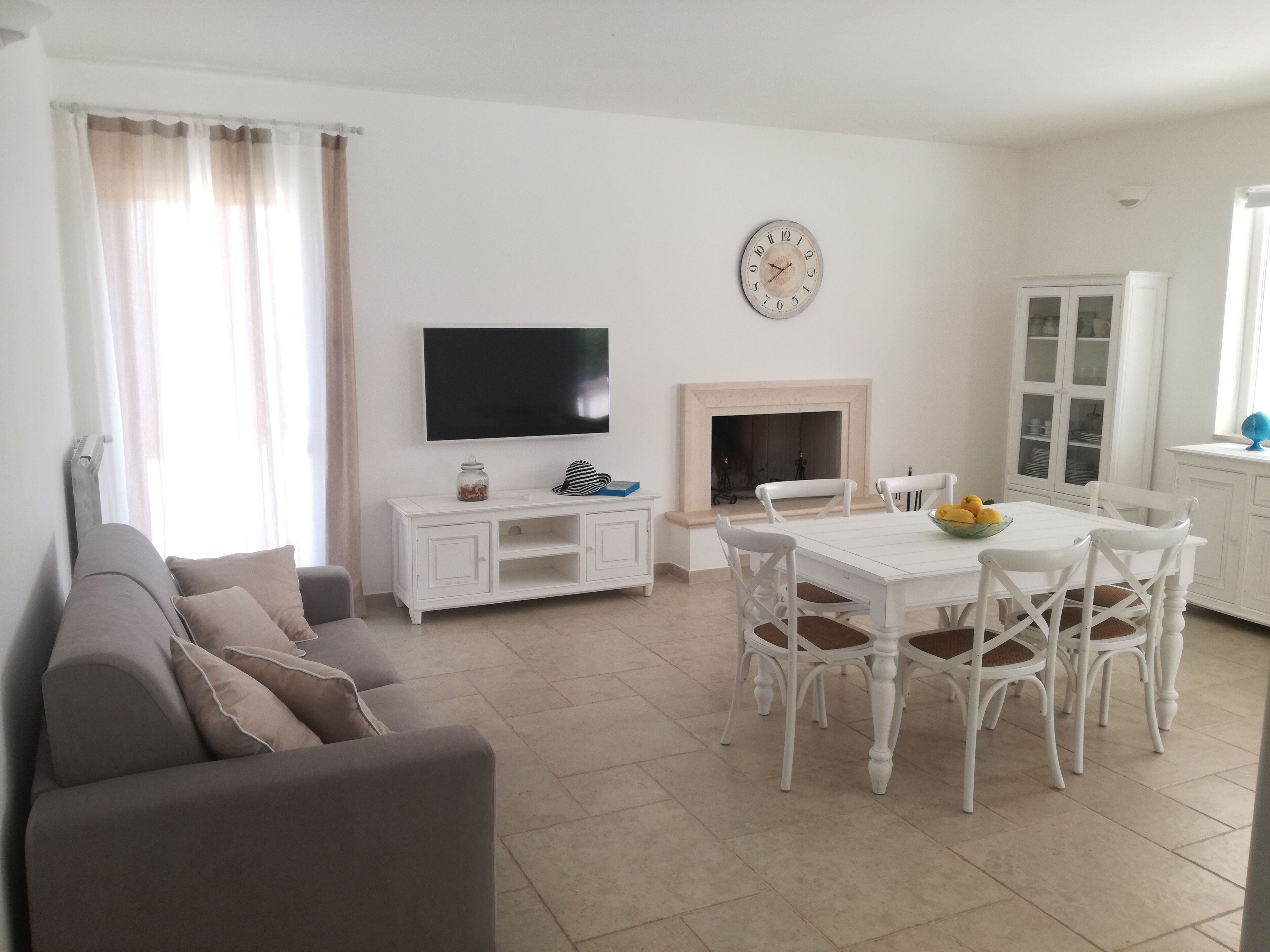 A living room with a sofa, a dining table with chairs, a flat-screen TV, a fireplace, and a clock on the wall.