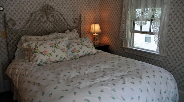 A bedroom with a floral-patterned bedspread, a decorative headboard, and a nightstand with a lamp. - Stone Throw Cottage (Bar Harbor)