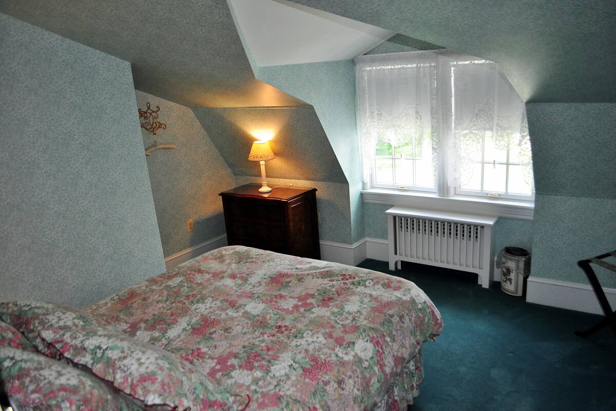 A bedroom with a bed, a nightstand with a lamp, a window with sheer curtains, and a radiator.