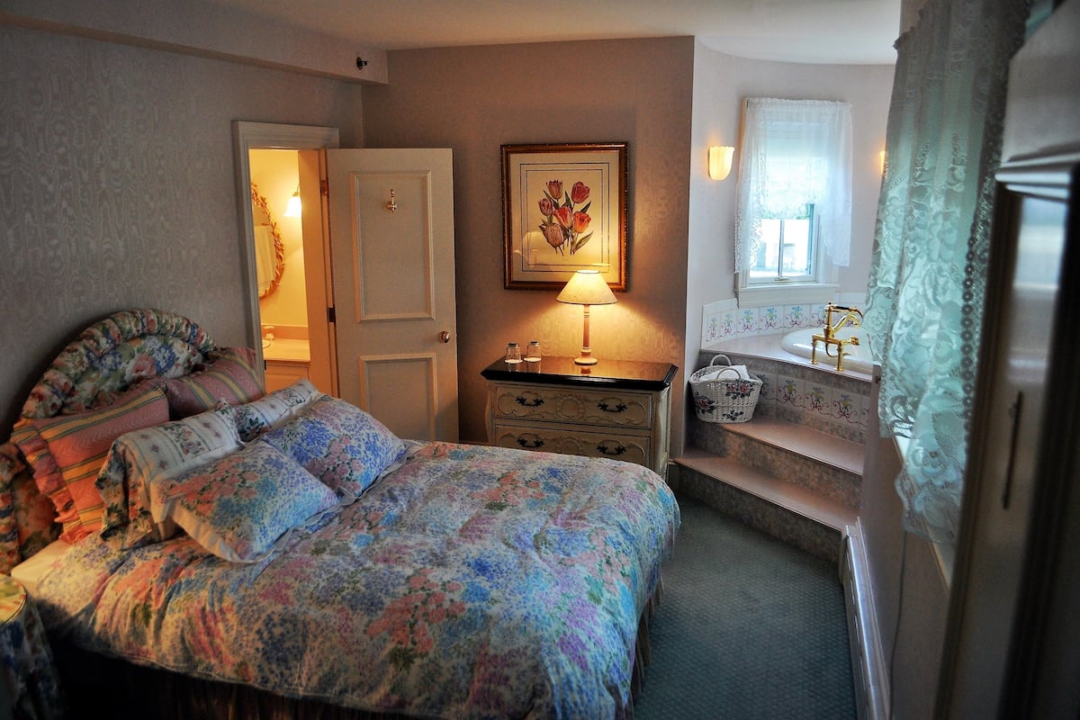 A bedroom with a floral bedspread, a bedside lamp, a dresser, and a window with curtains.