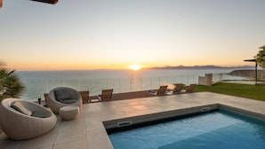 Zona junto a la piscina con sillones de descanso, una piscina pequeña y vista al océano al atardecer.