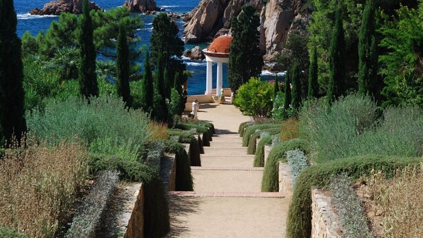 A pathway leading to a building with a red roof, surrounded by greenery and overlooking a coastal area.