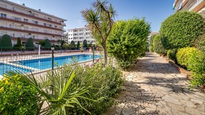 A pool area with a stone pathway, surrounded by greenery and a multi-story building in the background.