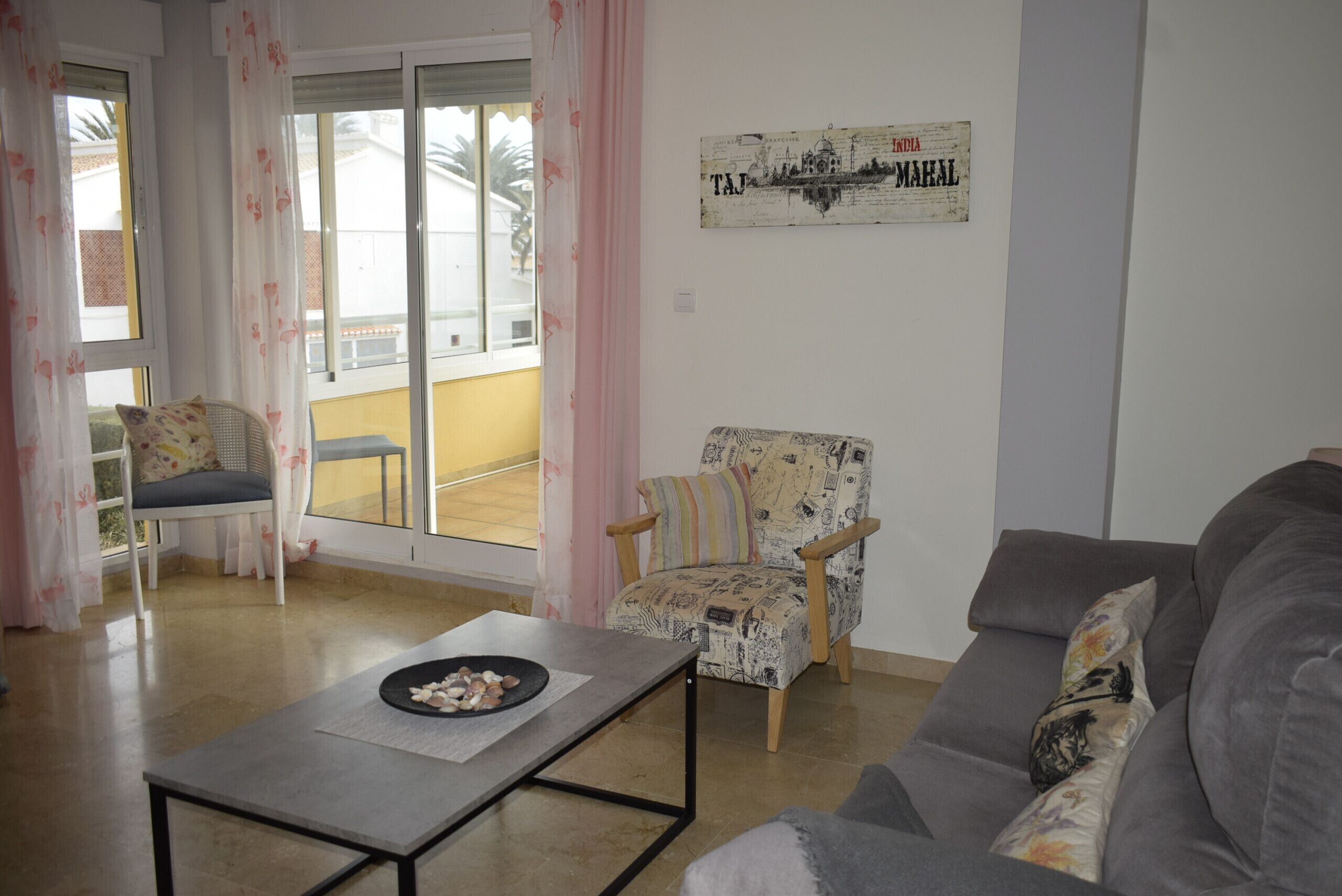Una sala de estar con un sofá gris, un sillón estampado, una mesita con un cuenco de aperitivos y una ventana con cortinas.