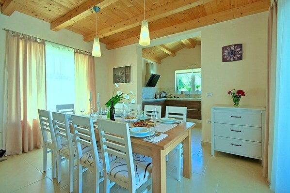 Un comedor con una mesa de madera dispuesta para cuatro, sillas blancas e iluminación colgante. Se ve una cocina al fondo con un reloj en la pared.