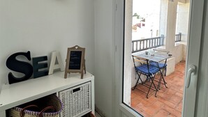 Una habitación con una puerta blanca que da a un balcón con mesa y sillas. Hay cestas de mimbre en el suelo y un pequeño estante con objetos decorativos.