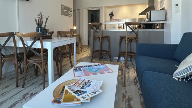 Un salón moderno con un sofá azul, una mesa de centro blanca con revistas, sillas de madera y una zona de cocina al fondo.